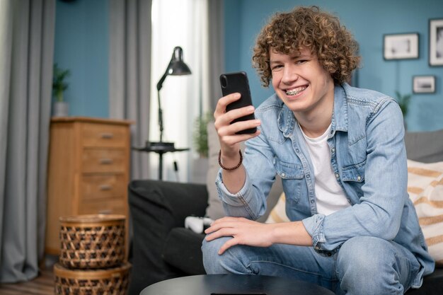 Portrait d'un adolescent caucasien aux cheveux bouclés dans un salon confortable à l'aide d'un smartphone à la maison