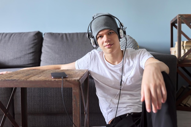 Portrait d'adolescent hipster au chapeau avec un casque assis sur le sol à la maison. Beau mec à la mode en regardant la caméra. Jeunesse, mode de vie et loisirs