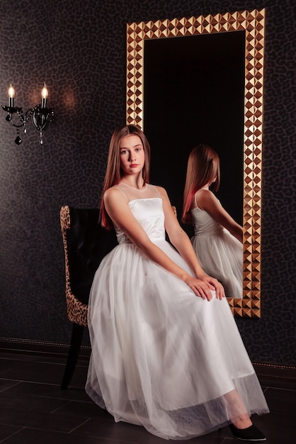Portrait d'une adolescente assez mignonne dans une robe élégante au miroir dans un intérieur sombre et élégant du salon. Émotions et pose des enfants. Concept de style, de mode et de beauté. Espace de copie pour le site