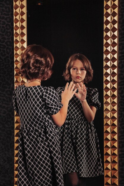 Portrait d'une adolescente assez mignonne vêtue d'une robe élégante au miroir dans un intérieur sombre et élégant du salon Émotions des enfants et pose Concept de style mode et beauté Espace de copie pour le site