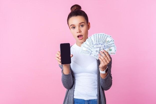 Portrait d'une adolescente brune étonnée avec une coiffure chignon dans des vêtements décontractés montrant un téléphone portable et des dollars avec un visage choqué surpris tourné en studio isolé sur fond rose