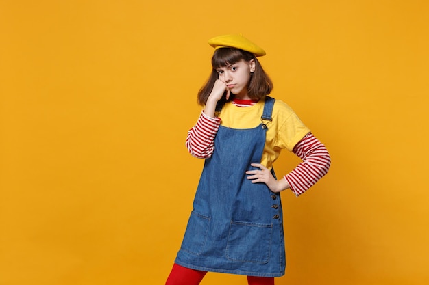 Portrait d'une adolescente concernée en béret français, robe d'été en jean mettant la main sur la joue isolée sur fond de mur jaune en studio. Émotions sincères des gens, concept de style de vie. Maquette de l'espace de copie.