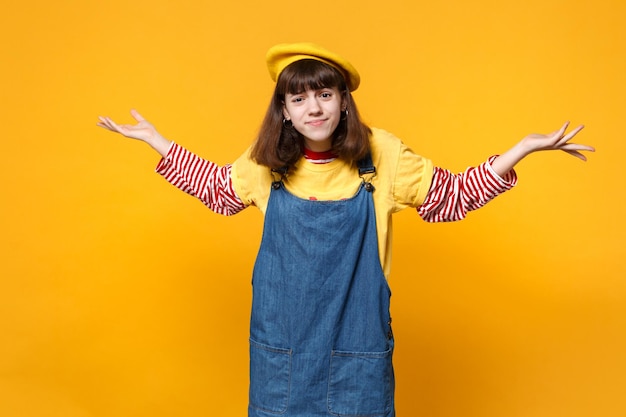 Portrait d'une adolescente découragée perplexe en béret français, robe d'été en denim écartant les mains isolées sur fond jaune en studio. Émotions sincères des gens, concept de style de vie. Maquette de l'espace de copie.