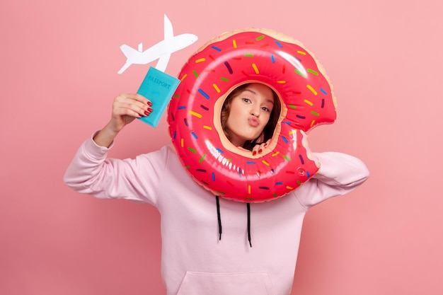 Photo portrait d'une adolescente drôle aux cheveux bouclés en sweat à capuche regardant à travers un anneau en caoutchouc beignet tenant un passeport avec un avion en papier à la main voyageant en studio intérieur isolé sur fond rose