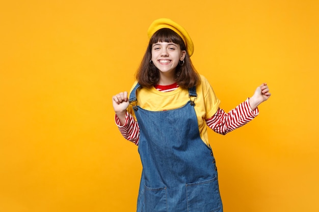 Portrait d'une adolescente drôle souriante en béret français tenant la main sur une robe d'été en denim isolée sur fond de mur jaune en studio. Émotions sincères des gens, concept de style de vie. Maquette de l'espace de copie.