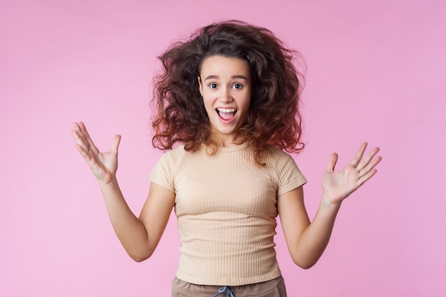 Portrait d'une adolescente folle et enthousiaste aux cheveux bruns bouclés échevelés volant vers le haut et dans des vêtements décontractés, levant les mains, criant de joie, submergée par l'excitation. tourné en studio, intérieur