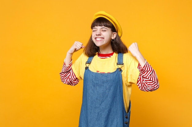 Portrait d'une adolescente heureuse en béret français, robe d'été en jean gardant les yeux fermés, faisant un geste gagnant isolé sur fond jaune. Émotions sincères des gens, concept de style de vie. Maquette de l'espace de copie.