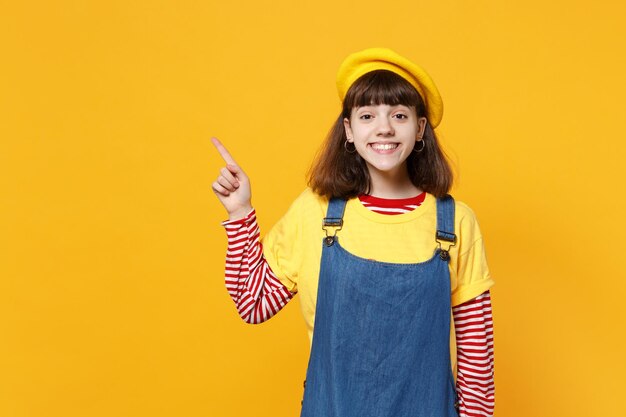 Portrait d'une adolescente joyeuse en béret français, robe d'été en jean pointant l'index de côté isolé sur fond jaune en studio. Émotions sincères des gens, concept de style de vie. Maquette de l'espace de copie.