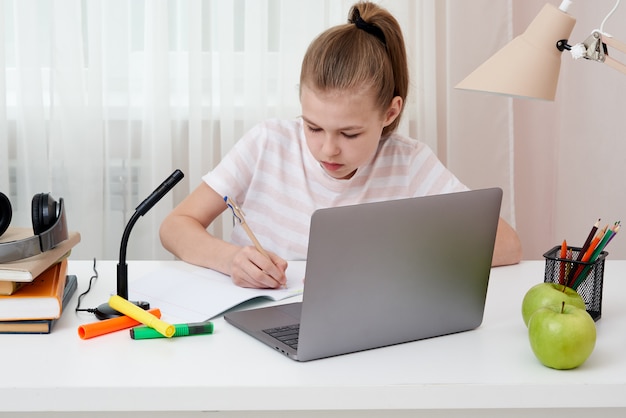 Portrait d'adolescente en ligne d'apprentissage avec des écouteurs et un ordinateur portable en prenant des notes dans un ordinateur portable assis à son bureau à la maison à faire leurs devoirs