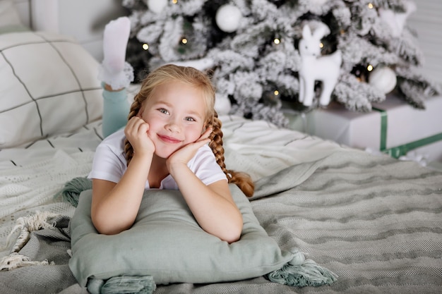 Portrait d'une adolescente avec des nattes allongée sur un lit avec des oreillers sur le fond d'un arbre de Noël. Mise au point sélective, arrière-plan flou.