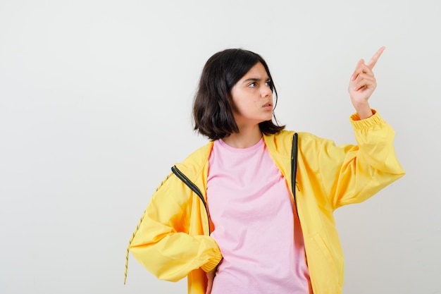 Portrait d'une adolescente pointant vers le coin supérieur droit en t-shirt, veste et à la vue de face perplexe