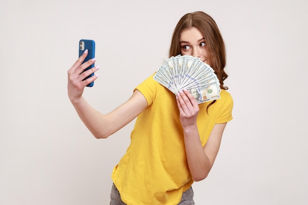 Portrait d'une adolescente riche aux cheveux bruns en T-shirt tenant de l'argent et prenant un selfie par téléphone portable, se vantant de gros bénéfices, de paris en ligne réussis. Tourné en studio intérieur isolé sur fond gris