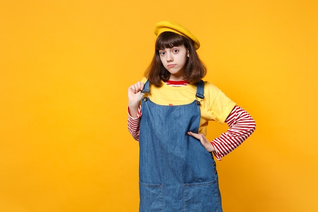 Portrait d'une adolescente sérieuse et lumineuse en béret français tenant la main sur une robe d'été en denim isolée sur fond de mur jaune en studio. Émotions sincères des gens, concept de style de vie. Maquette de l'espace de copie.