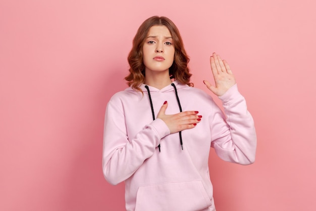 Portrait d'une adolescente sincère et honnête en sweat à capuche tenant la main vers le haut et sur la poitrine prêtant allégeance en prêtant serment avec une expression responsable Prise de vue en studio intérieure isolée sur fond rose