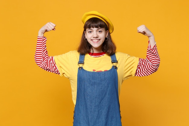 Portrait d'une adolescente souriante en béret français, robe d'été en denim montrant des biceps, des muscles isolés sur fond de mur jaune en studio. Émotions sincères des gens, concept de style de vie. Maquette de l'espace de copie.