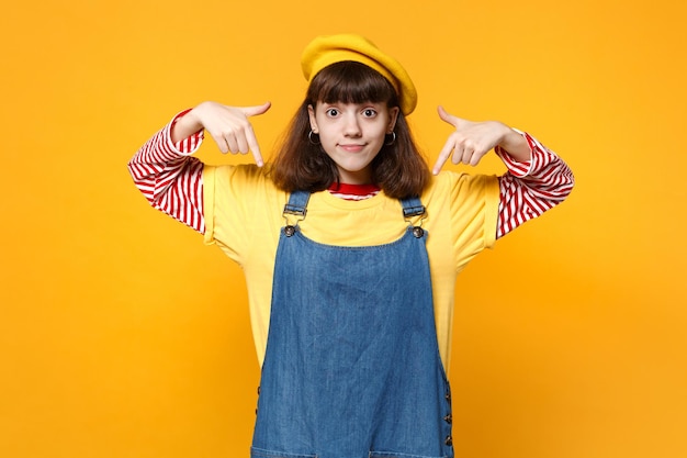 Portrait d'une adolescente souriante en béret français, robe d'été en denim pointant l'index vers le bas isolé sur fond jaune en studio. Émotions sincères des gens, concept de style de vie. Maquette de l'espace de copie.