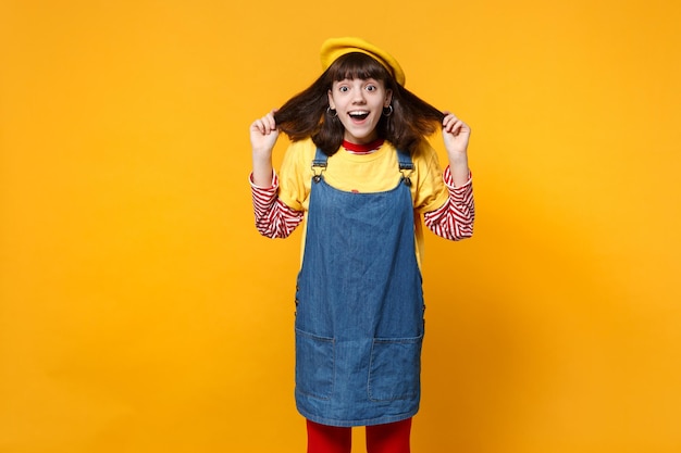 Portrait d'une adolescente surprise en béret français, robe d'été en jean tenant les cheveux, gardant la bouche ouverte isolée sur fond de mur jaune. Émotions sincères des gens, concept de style de vie. Maquette de l'espace de copie.