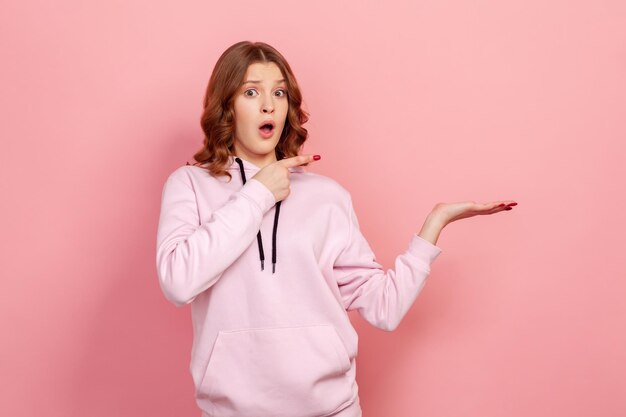 Portrait d'une adolescente surprise en sweat à capuche pointant le doigt et la paume vers l'espace vide, regardant la caméra avec la bouche ouverte et l'expression choquée. Studio intérieur tourné isolé sur fond rose