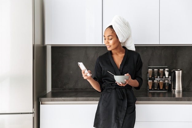 Portrait d'adorable femme afro-américaine portant une robe de chambre, à l'aide de téléphone portable dans la cuisine pendant le petit déjeuner