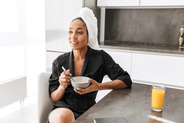 Portrait d'adorable femme afro-américaine portant une robe de chambre, prenant son petit déjeuner à la maison