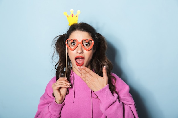 Portrait d'adorable fille princesse portant une couronne de papier en riant et tenant des lunettes de soleil drôles sur bâton sur bleu en studio