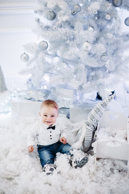 Portrait d'adorable petit bébé portant une chemise blanche et souriant à l'avant assis sur la neige artificielle près de l'arbre de Noël