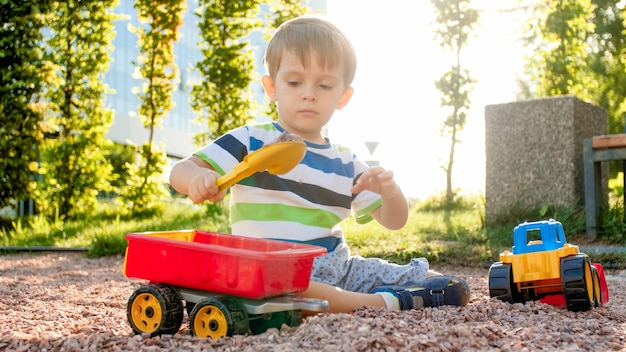 Portrait d'un adorable petit garçon de 3 ans jouant avec un camion jouet avec remorque sur l'aire de jeux du parc. Enfant creusant et construisant du sable