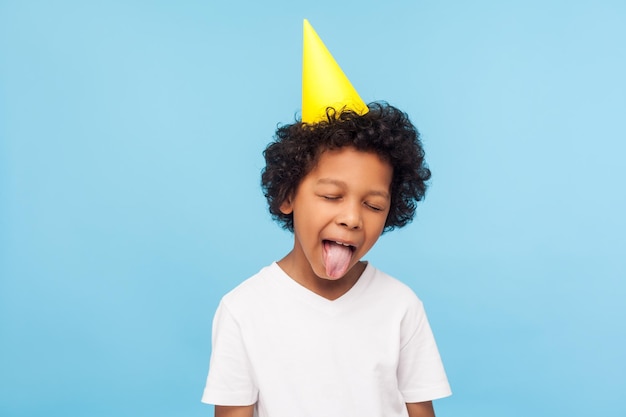 Portrait d'adorable petit garçon désobéissant drôle avec un cône de fête sur la tête qui sort la langue et garde les yeux fermés, un enfant méchant grimaçant s'amusant le jour de son anniversaire. tourné en studio, fond bleu