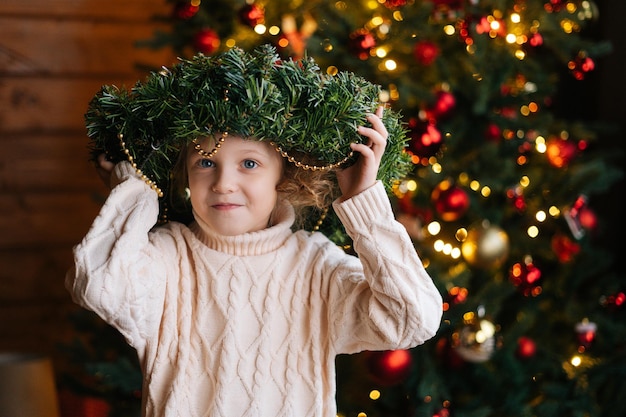 Portrait d'adorable petite fille blonde bouclée tenant Noël debout dans une pièce sombre et confortable avec un intérieur festif