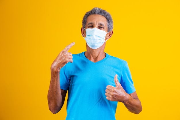 Portrait d'un afro âgé avec un masque médical chirurgical debout en chemise bleue avec le pouce en l'air, le pouce levé tout en pointant le masque sur son visage.