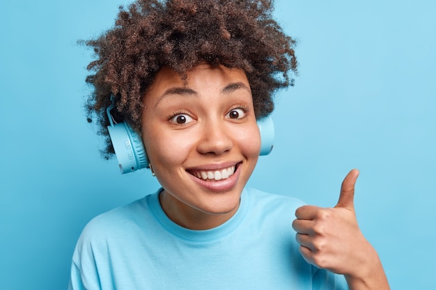 Portrait d'un Afro-américain aux cheveux bouclés écoute de la musique et aime sa piste préférée via un casque garde le pouce levé montre d'excellents sourires de signe heureusement isolés sur le mur bleu. Concept de langage corporel