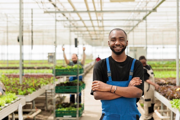 Portrait d'un afro-américain souriant dans une serre moderne avec des travailleurs préparant des caisses d'aliments biologiques pour la livraison. Agriculteur de cultures bio posant heureux dans un environnement hydroponique pour la culture de cultures vertes.