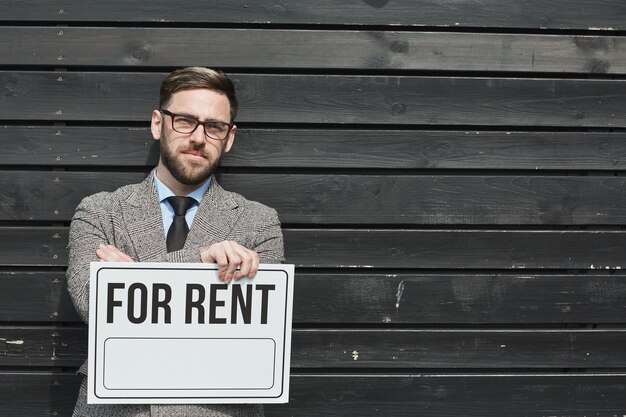 Portrait D'agent Immobilier Barbu Mature à Lunettes Tenant Une Pancarte à Louer Contre Le Mur Noir à L'extérieur