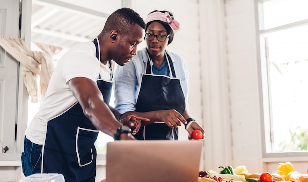 Portrait d'amour couple afro-américain s'amusant à cuisiner ensemble et à la recherche de recette sur Internet avec ordinateur portable pour préparer le délicieux manger dans la cuisine à la maison