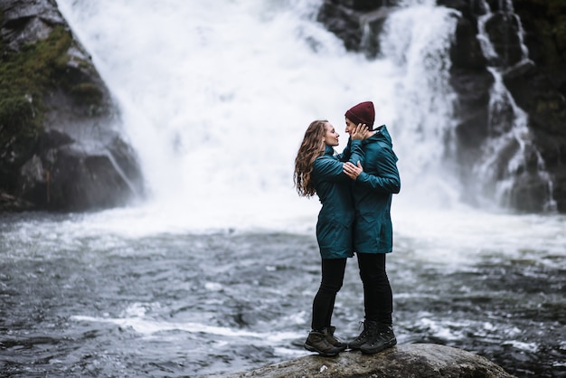 Portrait d'amoureux en imperméables verts, debout sur une pierre, contre une cascade