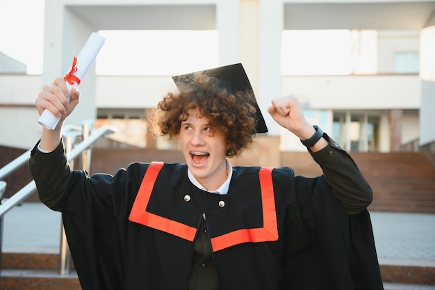 Portrait en angle bas d'un heureux diplômé masculin triomphant debout près de l'université tenant un diplôme