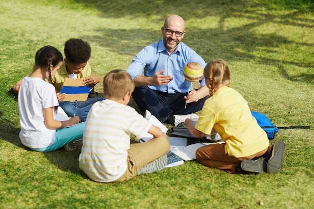 Portrait d'angle élevé de professeur de sexe masculin chauve pointant sur le modèle de planète et souriant tout en parlant à un groupe d'enfants pendant les cours en plein air au soleil