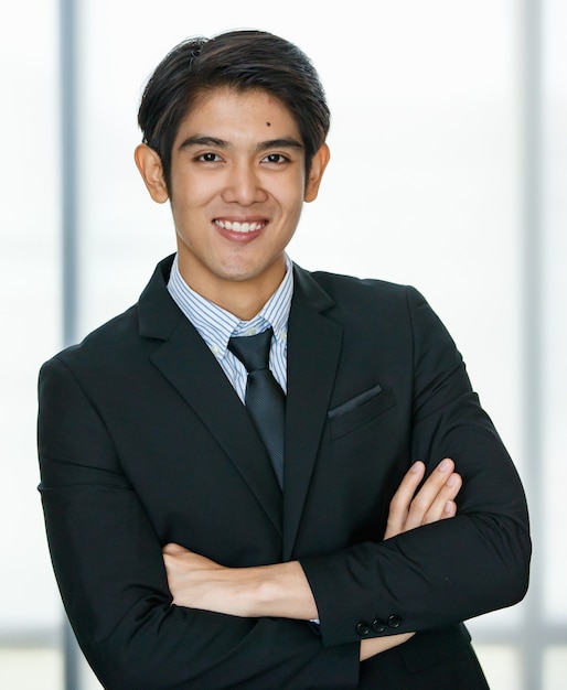 Photo portrait des années 20, beau jeune homme d'affaires asiatique charismatique portant un costume formel avec une cravate, souriant avec confiance et intelligemment, croisant les bras avec positivité, confiance, succès dans un bureau intérieur.