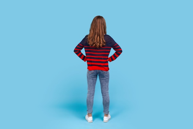 Photo portrait arrière de pleine longueur d'une femme portant un pull rayé de style décontracté posant à l'envers avec les mains sur les hanches debout calme méconnaissable en attente prise de vue en studio intérieur isolé sur fond bleu