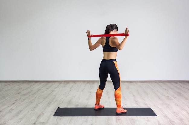 Photo portrait arrière pleine longueur d'une femme sportive portant un haut noir et des leggings orange effectue des exercices pour les muscles du dos et des mains, entraînement avec bande de résistance sur fond blanc. intérieur