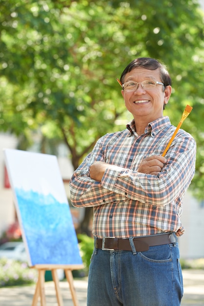 Portrait d'un artiste asiatique mature souriant avec un crayon derrière l'oreille debout avec un pinceau dans le parc de la ville