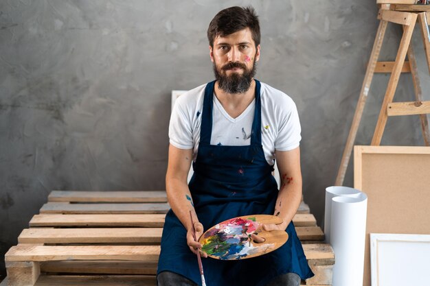 Portrait d'un artiste caucasien barbu assis dans un studio d'art regardant la caméra et souriant tenant une palette avec des peintures et un pinceau