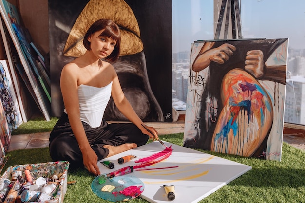 Portrait d'une artiste féminine assise sur un tapis dans un studio d'art avec vue sur la ville parmi les portraits et l'équipement de peinture