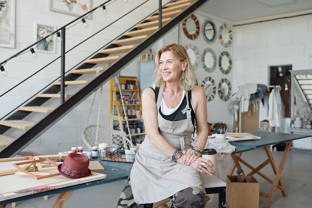 Portrait d'un artiste mature souriant assis dans un studio d'art avec des photos et buvant du café