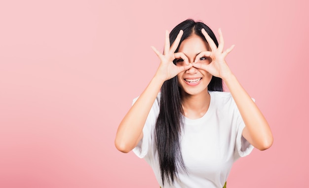 Portrait Asiatique belle jeune femme debout, Elle a fait signe du doigt OK symbole pour s'accorder près des yeux en regardant la caméra, prise de vue en studio isolée sur fond rose, Femme thaïlandaise d'accord main signe copie espace