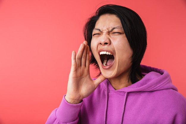 Portrait d'un asiatique en colère portant un sweat-shirt criant tout en étant furieux isolé sur un mur rouge