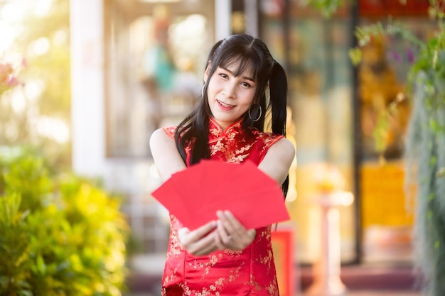 Portrait asiatique femme transgenre portant une décoration traditionnelle chinoise cheongsam rouge tenant des enveloppes rouges à la main au sanctuaire chinois pour le Nouvel An chinois