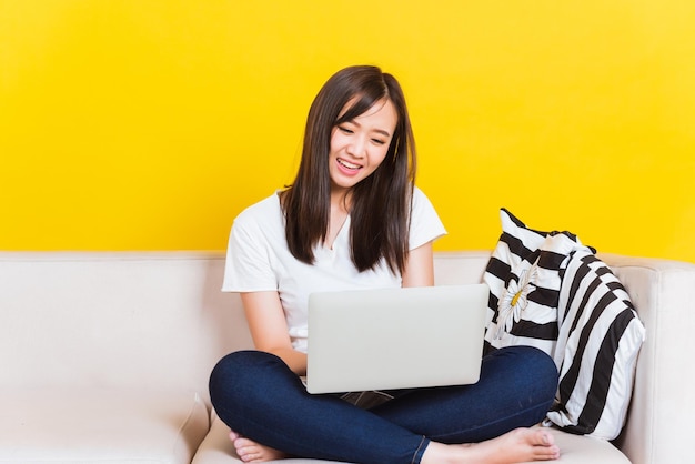 Portrait asiatique d'heureuse belle jeune femme travaille à la maison, elle est assise sur un canapé à l'aide d'un ordinateur portable dans la maison salon studio tourné isolé sur fond jaune