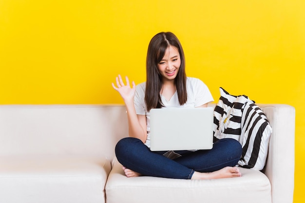 Portrait asiatique d'heureuse belle jeune femme travaille à la maison, elle est assise sur un canapé, lève la main et dit bonjour sur un ordinateur portable dans la maison salon studio tourné isolé sur fond jaune