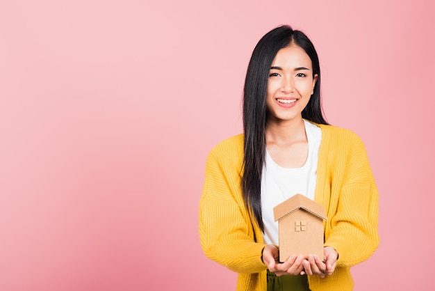 Portrait asiatique heureux belle jolie jeune femme excitée souriante tenant le modèle de maison à portée de main, prise de vue en studio isolée sur fond rose, femme courtier détenant l'assurance immobilière à domicile et le concept bancaire